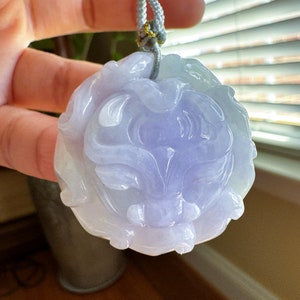 May include: A lavender jade pendant carved in the shape of a lion's head. The pendant is attached to a gray cord.
