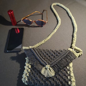 Può includere: Una borsa a tracolla in crochet nero e verde con un dettaglio a frangia e una lunga tracolla. La borsa è appoggiata su una superficie scura con un paio di occhiali da sole, un telefono cellulare e un tubetto di rossetto rosso.