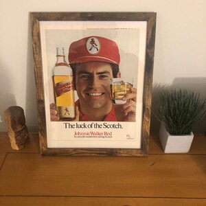 May include: A framed vintage advertisement for Johnnie Walker Red Scotch whisky. The ad features a man wearing a red cap and a red shirt, holding a bottle of Johnnie Walker Red and a glass of scotch. The text on the ad reads "The luck of the Scotch. Johnnie Walker Red. So smooth-world's best selling Scotch."