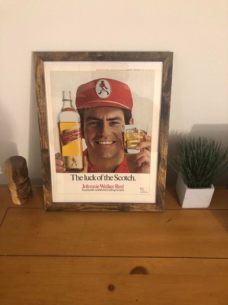 May include: A framed vintage advertisement for Johnnie Walker Red Scotch whisky. The ad features a man wearing a red cap and a red shirt, holding a bottle of Johnnie Walker Red and a glass of scotch. The text on the ad reads "The luck of the Scotch. Johnnie Walker Red. So smooth-world's best selling Scotch."