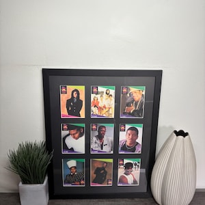 May include: A framed collection of nine vintage trading cards featuring portraits of musicians. The cards are arranged in a black frame, with a white border. The frame is displayed on a wooden surface, next to a decorative vase and a small potted plant.
