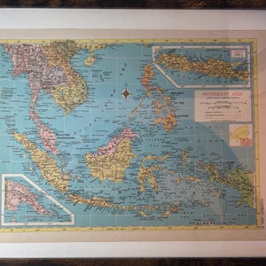 May include: A framed vintage map of Southeast Asia, featuring countries in shades of yellow, pink, and green against a blue ocean. The map includes the text "SOUTHEAST ASIA" and is encased in a dark wooden frame.