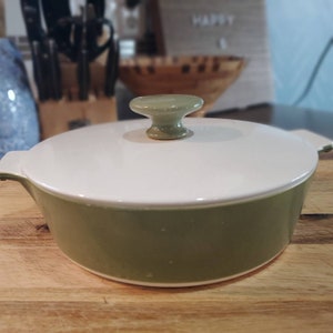 May include: A green ceramic casserole dish with a white lid and a knob on top. The dish is sitting on a wooden surface.