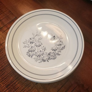 May include: A white ceramic dinner plate with a blue and white floral design. The plate has a thin, dark blue rim with a subtle pattern.
