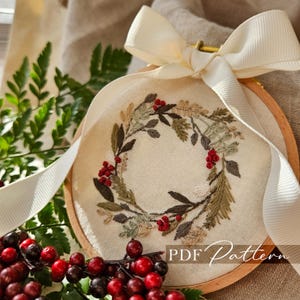 May include: An embroidered wreath with red berries and green and brown leaves is displayed in a wooden hoop. A cream-colored ribbon is tied in a bow at the top. The text "PDF Pattern" is visible.