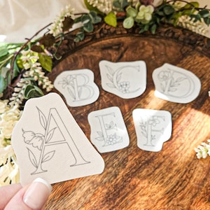 May include: A hand holds a cream-colored patch with the letter "A" and floral design. Other patches with letters B, C, E, and F, also with floral designs, are arranged on a wooden surface. The patches are outlined in gray.