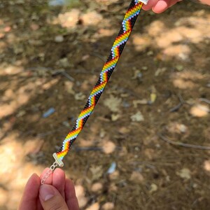 May include: A beaded bracelet with a rainbow chevron pattern. The bracelet features a black background with zig-zagging lines of red, orange, yellow, green, blue, and purple beads. The bracelet is held by a silver clasp.