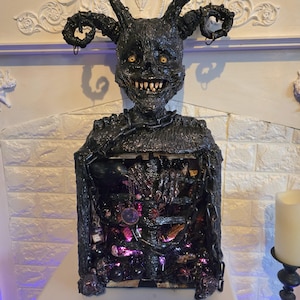 May include: A black, horned sculpture of a demonic figure with a wide grin and yellow eyes. The figure has a chain around its neck and a chest cavity filled with glowing purple and pink lights and decorative elements. The sculpture is on a white surface.