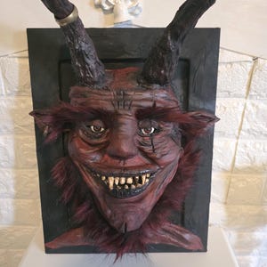 May include: A detailed sculpture of a demonic face. The face is a deep red with dark horns, a wide, toothy grin, and red fur around the neck. The sculpture is mounted on a black rectangular frame.