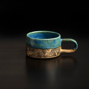 Puede incluir: Taza de cerámica hecha a mano con parte superior esmaltada en azul y verde y base texturizada de color beige. La taza presenta un asa curvada y un diseño artístico único. Ideal para café o té.