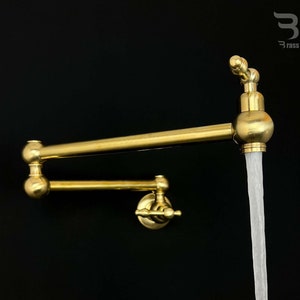 May include: A gold-toned kitchen faucet with a swivel spout and a wall-mounted design. The faucet is turned on and water is flowing from the spout.