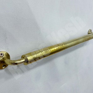 Könnte beinhalten: Eine goldene, gehämmerte Messing-Handtuchhalterung mit einem gebogenen Arm und einer runden Wandhalterung. Die Stange ist etwa 45 cm lang.