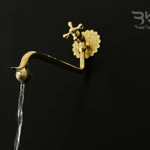 May include: A gold-toned brass faucet with a decorative floral design and a cross-shaped handle. Water is flowing from the spout.