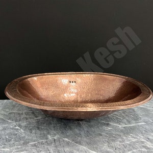 Puede incluir: Un lavabo de baño ovalado de cobre martillado con un borde decorativo.