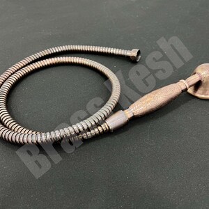 Pu&ograve; includere: Un set doccia color bronzo composto da soffione e tubo flessibile. Il soffione ha una forma a campana e un manico ornato. Il tubo flessibile &egrave; di colore argento metallizzato. Il set &egrave; esposto su una superficie scura.