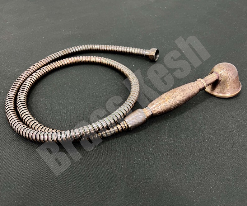 Pu&ograve; includere: Un set doccia color bronzo composto da soffione e tubo flessibile. Il soffione ha una forma a campana e un manico ornato. Il tubo flessibile &egrave; di colore argento metallizzato. Il set &egrave; esposto su una superficie scura.
