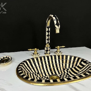 Può includere: Un lavabo da bagno ovale a strisce bianche e nere con un bordo in ottone e un rubinetto abbinato. Il rubinetto ha una maniglia con motivo bianco e nero.