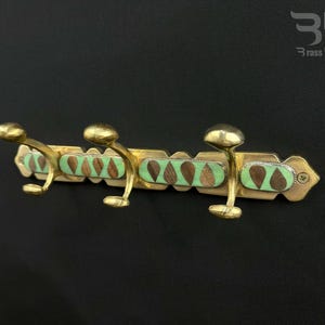May include: Brass coat rack with three hooks and a decorative green and brown inlay. The rack is mounted on a black wall.