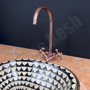 May include: A black and white mosaic sink with a copper tap. The sink is oval shaped and has a geometric pattern. The tap has a hammered finish and two cross-shaped handles.