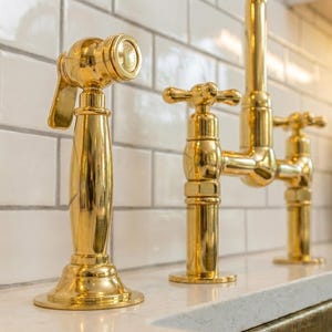 May include: Close-up of a gold-colored kitchen faucet set. The set includes a sprayer, a faucet with cross handles, and a tall faucet. The fixtures are set against a white tile backsplash.
