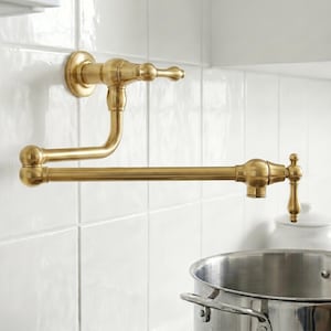 May include: A wall-mounted, brass-colored pot filler faucet with a classic design. The faucet features a curved spout and a handle for water control, mounted on a white tiled wall. A stainless steel pot is below the faucet.