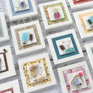 May include: A selection of framed art pieces with bird motifs. Each square frame is white, with a central panel displaying a bird design. The birds are in various colours, and the frames have decorative borders. The artwork is presented on a light wooden surface.