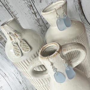 May include: Three pairs of earrings displayed on white ceramic vases. One pair features leaf-shaped pendants with a textured design. Another pair has light blue, teardrop-shaped pendants. The third pair has light blue, oval-shaped pendants. All earrings have silver-colored hooks.