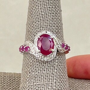 May include: A silver ring with a large oval ruby surrounded by smaller diamonds. The band is set with smaller rubies.