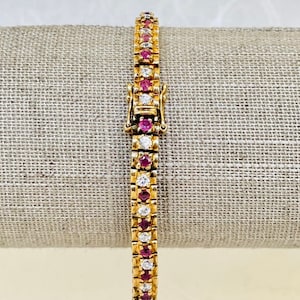 Könnte beinhalten: Ein goldenes Tennisarmband mit abwechselnd rosafarbenen und weißen Edelsteinen. Das Armband hat einen Verschluss.