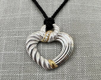 Puede incluir: Un collar con un colgante en forma de corazón de plata y oro, con un diseño de cuerda y una fila de piedras transparentes. El collar está en un cordón negro.