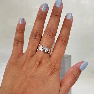 May include: A silver ring with a clear stone set in a curved band. The ring is worn on the middle finger of a hand with light blue nail polish.