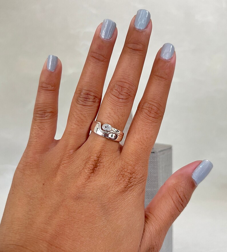 May include: A silver ring with a clear stone set in a curved band. The ring is worn on the middle finger of a hand with light blue nail polish.