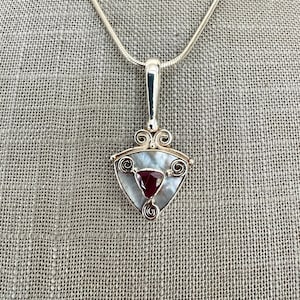 May include: A silver pendant necklace with a triangular shaped charm featuring a red gemstone and a white mother of pearl background. The charm is surrounded by intricate silver scrollwork.