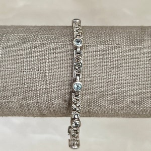 May include: A silver bracelet with a delicate chain and small blue gemstones. The bracelet is adorned with intricate marquetry details.