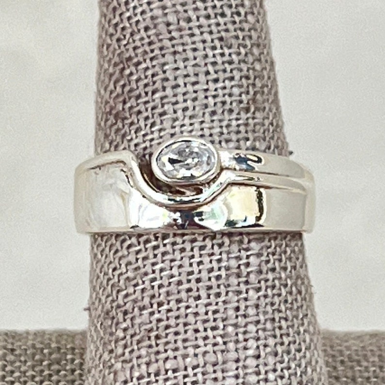 May include: A silver ring with a wave design and a clear oval-shaped stone.
