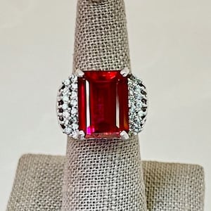 May include: A silver ring with a large, red, emerald-cut gemstone surrounded by smaller, clear gemstones.