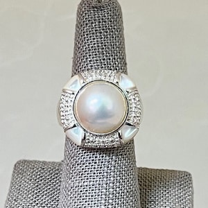 Könnte beinhalten: Ein silberner Ring mit einem großen, runden, irisierenden Perlmutt-Mittelstein. Der Perlmutt wird von einer quadratischen Fassung mit weißen Akzenten und kleinen, funkelnden Steinen umgeben. Der Ring wird auf einem grauen Ringhalter präsentiert.
