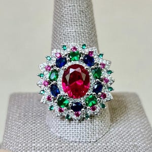 May include: A silver ring featuring a large red gemstone surrounded by smaller green, blue, and pink gemstones.