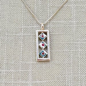 May include: A silver necklace with a rectangular pendant featuring three red gemstones set in a geometric pattern.