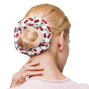 Op de afbeelding: Een witte scrunchie met een rode kersenprint. De scrunchie is gemaakt van stof en is om het haar van een vrouw gebonden.