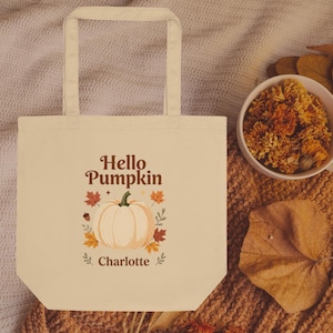 Könnte beinhalten: Eine cremefarbene Stofftasche mit einer zentralen Grafik eines hellorangen Kürbisses, umgeben von bunten Herbstblättern und Eicheln. Der Text "Hello Pumpkin" ist über dem Kürbis und der Name "Charlotte" unter dem Kürbis aufgedruckt.