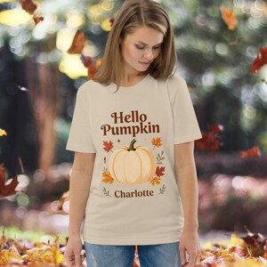 Peut inclure: Un t-shirt beige à manches courtes avec le texte marron Hello Pumpkin au-dessus d'une grande illustration de citrouille orange clair entourée de petites feuilles d'automne orange et marron, avec le nom Charlotte imprimé en marron.