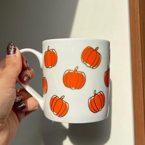 Könnte beinhalten: Weiße Keramik-Tasse mit orangefarbenen, goldumrandeten Kürbissen. Die Tasse hat einen gebogenen Henkel. Es ist ein saisonaler Artikel, perfekt für den Herbst. Die Tasse wird von einer Hand gehalten.
