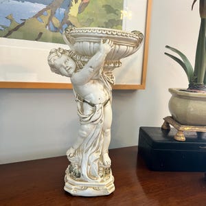 May include: A cream-colored cherub figurine holding a decorative bowl. The cherub is sculpted with detailed features and gold accents. The base of the figurine has an ornate design. The figurine is on a dark brown surface.