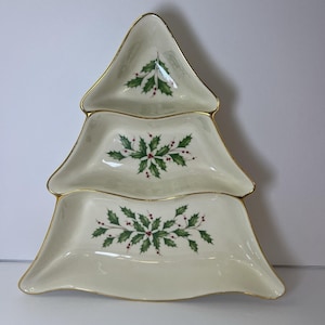 May include: A tiered serving dish in the shape of a Christmas tree. The off-white porcelain dish has a gold trim and is decorated with green holly leaves and red berries. The tree-shaped dish is perfect for serving snacks or appetizers during the holidays.