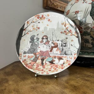 May include: A decorative ceramic plate featuring a whimsical illustration of dogs in a park setting. The scene includes a woman reading a newspaper with dogs on a bench and on the ground, with autumn leaves and a city skyline in the background.