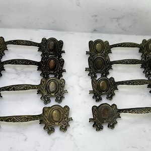 May include: Eight antique bronze drawer pulls with ornate floral and oval designs. Each pull has a central oval medallion and decorative flourishes. The pulls are arranged in two sets of four, showcasing their vintage aesthetic.