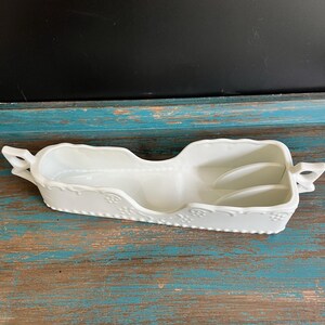 May include: A white ceramic serving dish shaped like a sleigh with a long, narrow body, two handles on the ends, and a scalloped rim. The exterior features a raised beaded texture and floral relief patterns along the base and sides.