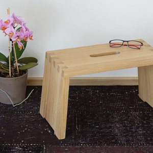 Könnte beinhalten: Ein heller Holzhocker mit einer rechteckigen Oberseite und einem kleinen Ausschnitt in der Mitte. Der Hocker steht auf einem dunkelbraunen Teppich mit einer rosa Orchideenpflanze in einem braunen Topf links.