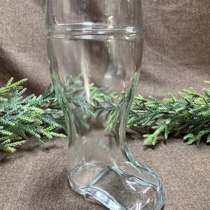 May include: A clear glass boot-shaped vase. The vase has a wide top and tapers down to a foot shape. The glass is transparent, allowing a view of the background. The vase is likely for decorative purposes.